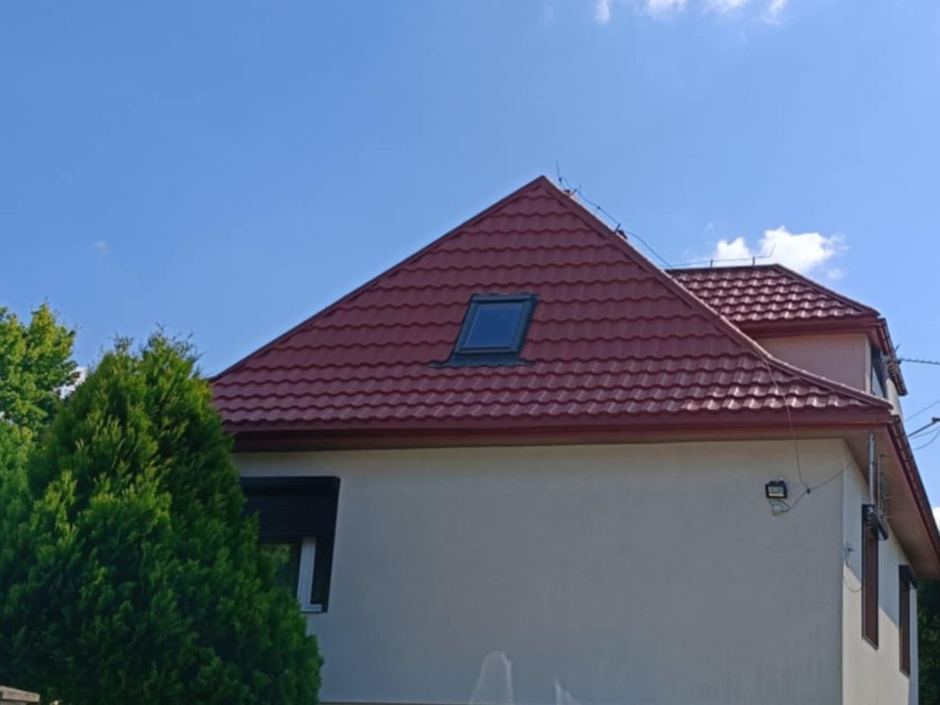 Dom z czerwonym dachem z blachodachówki trapezowej, widok z dołu, okno dachowe, orynnowanie, błękitne niebo, drzewo iglaste z lewej strony.