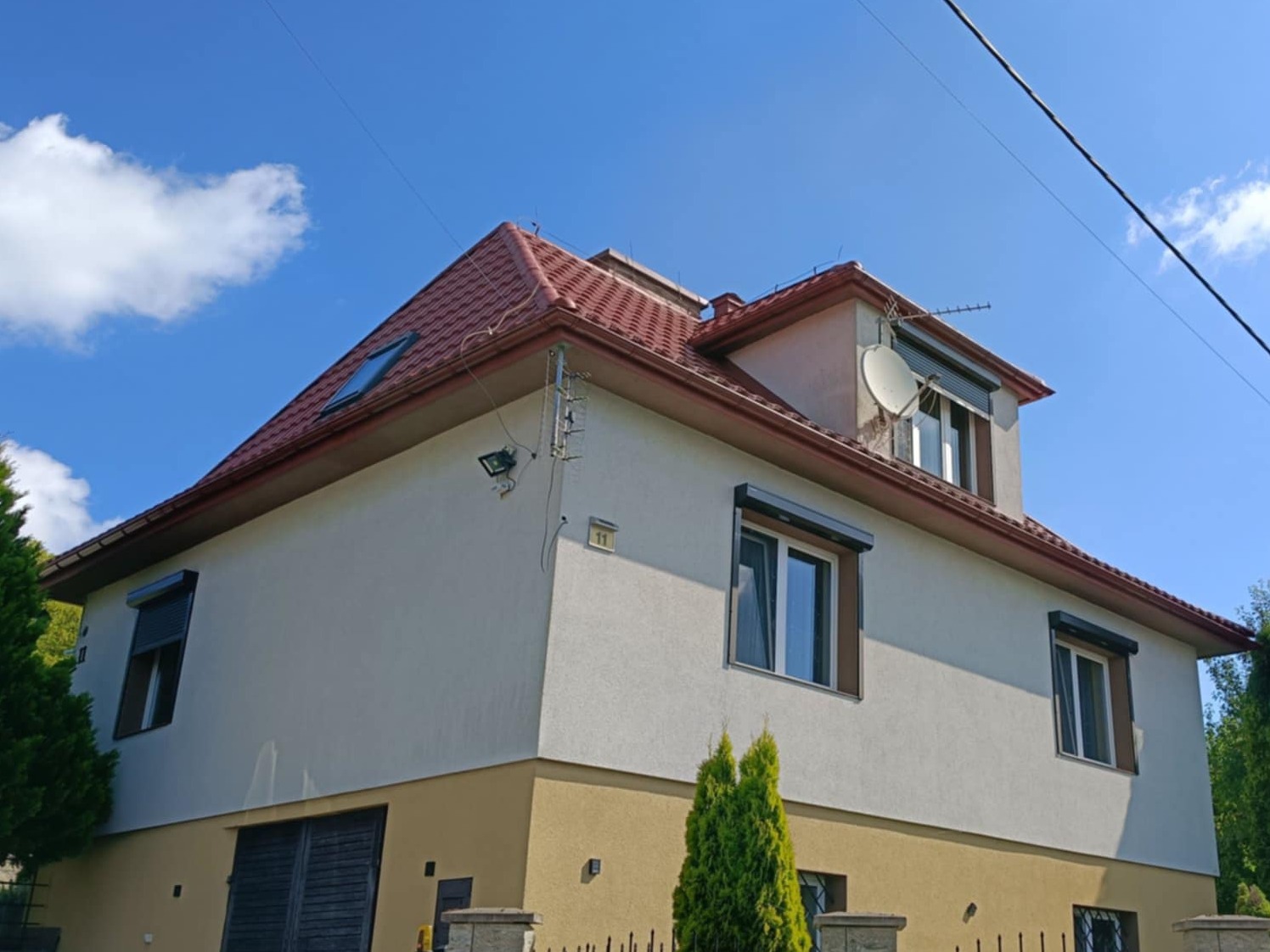 Dom z czerwonym dachem, widok z dołu na elewację i dach z oknem dachowym. Zamknięte rolety zewnętrzne w oknach. Antena satelitarna na dachu. Słoneczny dzień.