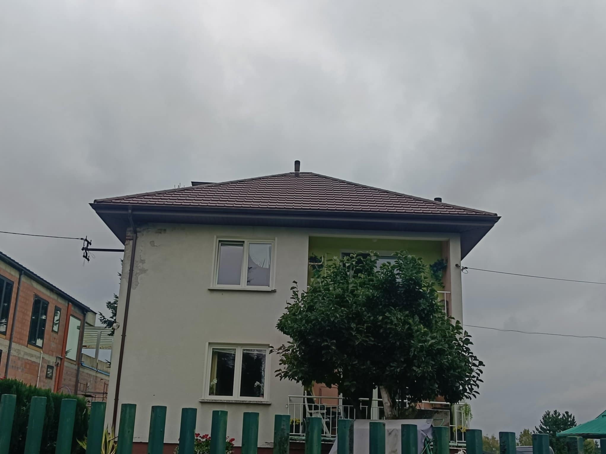 Dom z brązowym dachem widziany spod niskiego kąta, z zielonym ogrodzeniem na pierwszym planie i pochmurnym niebem w tle. Widoczny fragment budynku w budowie obok.
