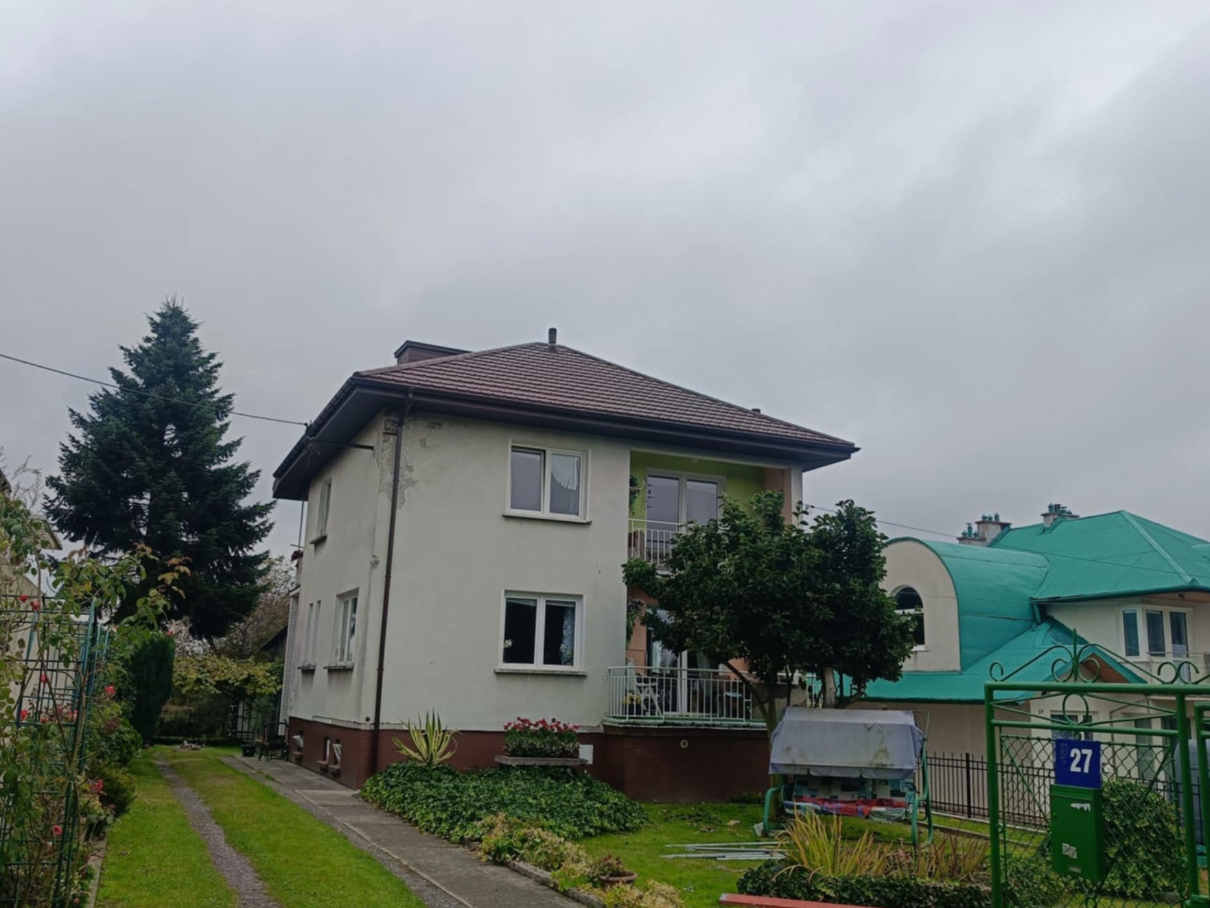 Dwupiętrowy dom z brązowym dachem i zielonym elementem balkonu, otoczony zielenią. W tle widoczny dom z zielonym dachem. Pochmurne niebo.