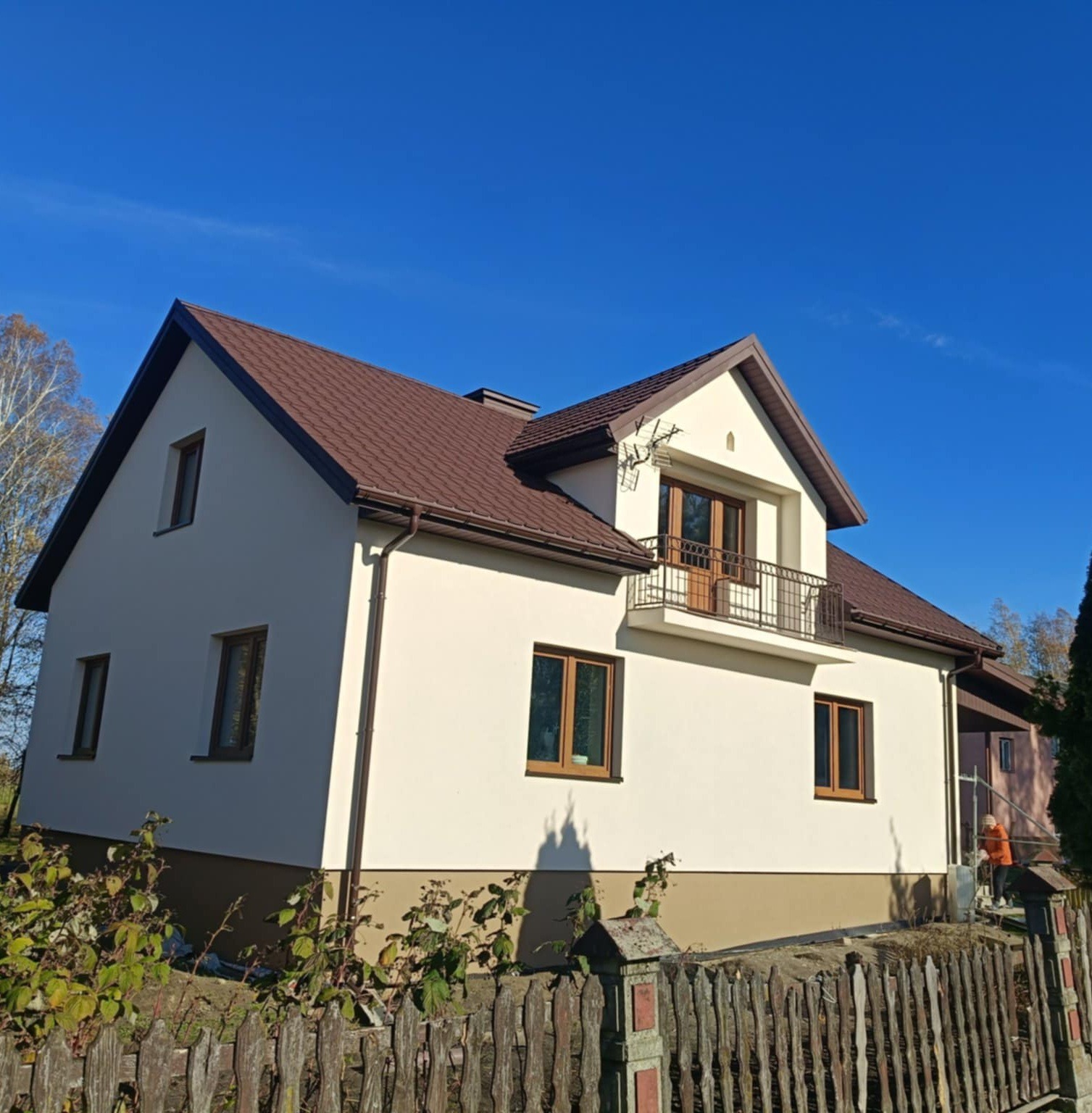 Dom jednorodzinny z brązowym dachem i rynnami, kremowa elewacja, balkon z metalową balustradą. Widok z dołu, ogrodzenie drewniane na pierwszym planie, błękitne niebo.