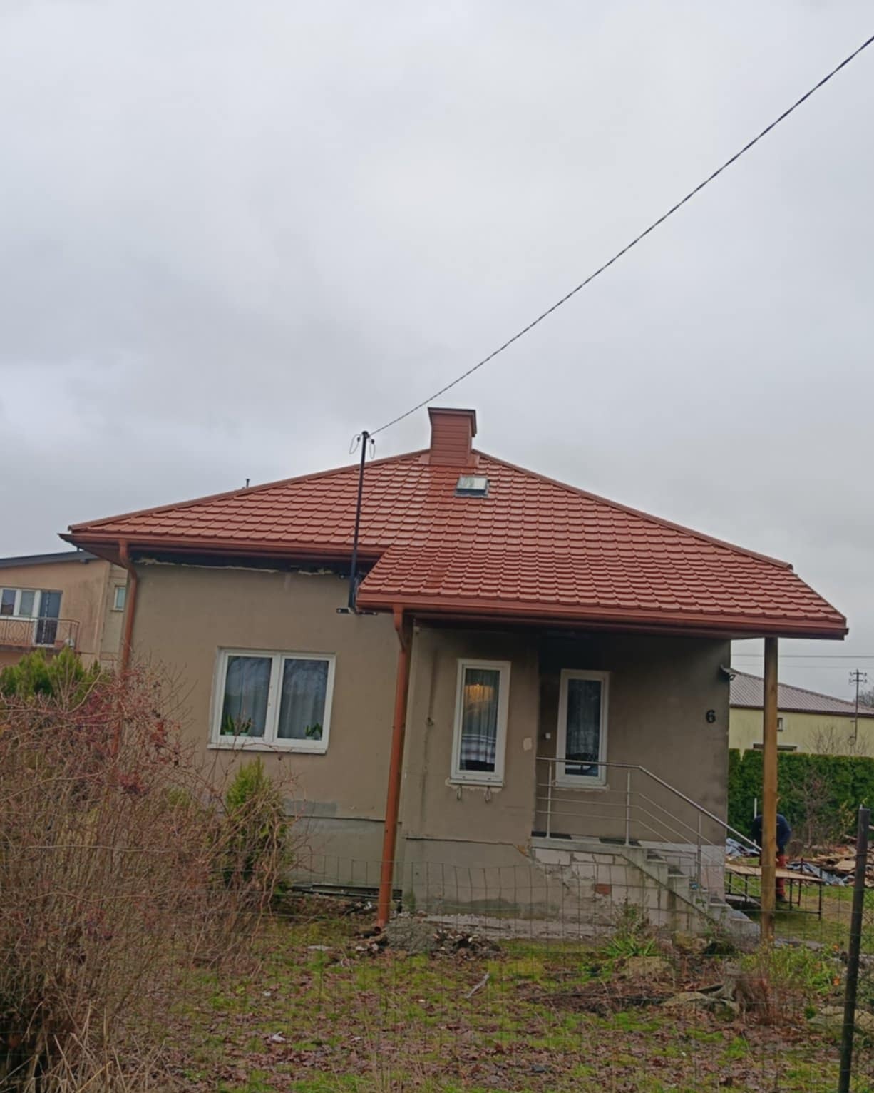 Dom jednorodzinny z nowym, czerwonym dachem z dachówki ceramicznej. Widoczne rynny w kolorze dachu i komin. Pochmurne niebo w tle. Ujęcie z poziomu gruntu.