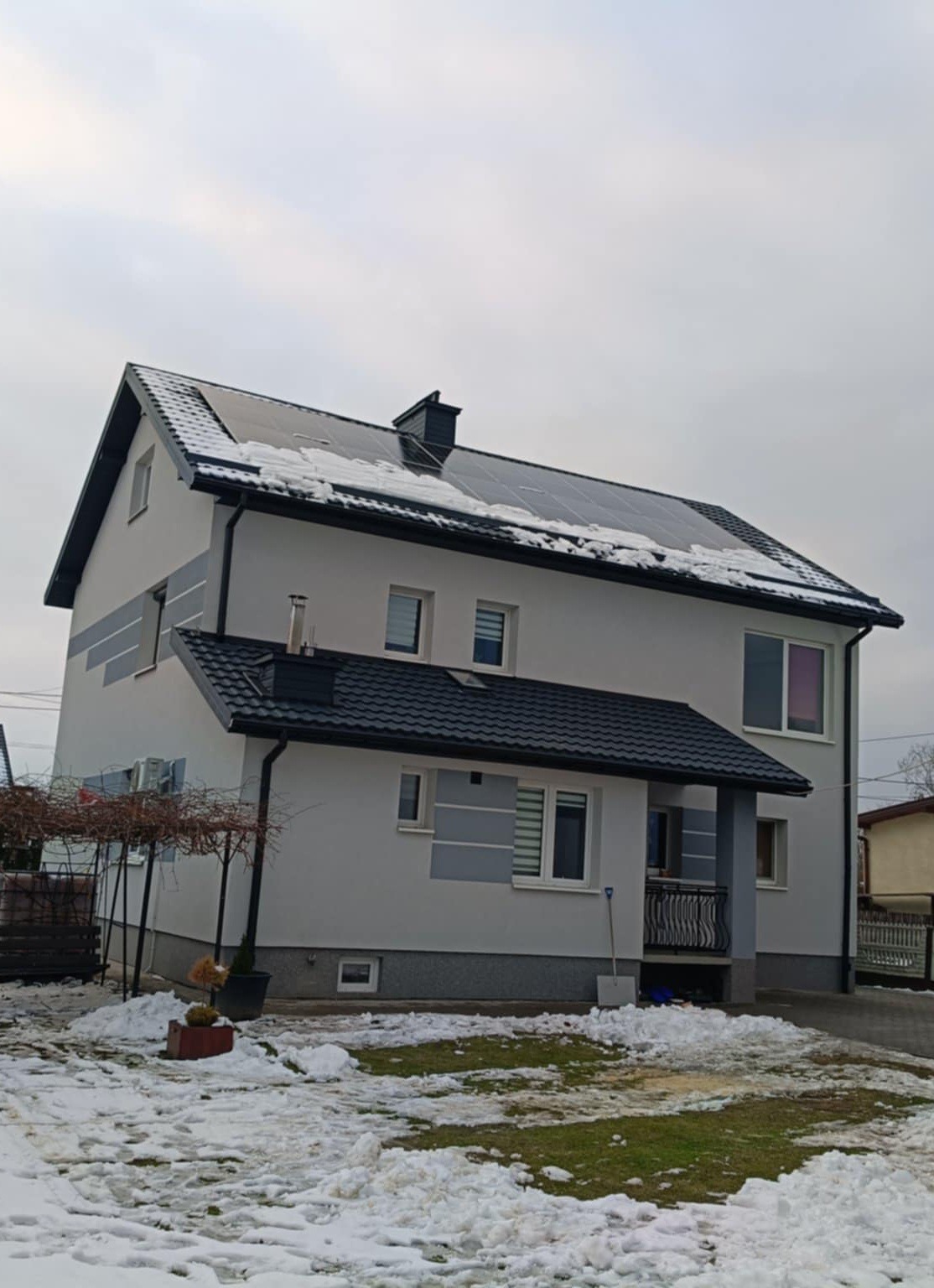Dwupiętrowy dom z ciemną dachówką i panelami słonecznymi, częściowo pokryty śniegiem. Widoczne rynny i obróbki blacharskie. Zima.