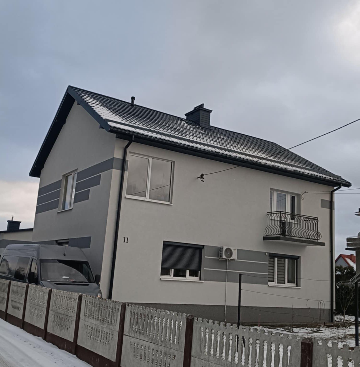 Dwukondygnacyjny dom z szarym dachem i elewacją w odcieniach szarości, widoczny zza betonowego ogrodzenia w zimowej scenerii. Na dachu leży cienka warstwa śniegu.