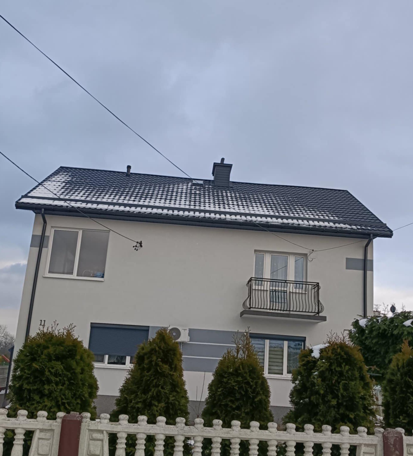 Dom jednorodzinny z ciemnoszarym dachem pokrytym śniegiem, widoczny komin i rynny. Biała elewacja z szarymi pasami, balkon z czarną balustradą, tuje wzdłuż ogrodzenia.