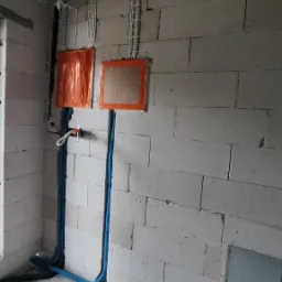 Surowe wnętrze w trakcie prac: widoczne kable elektryczne prowadzone po ścianach z bloczków, zabezpieczone puszki instalacyjne oraz rury karbowane w kolorze niebieskim.