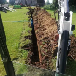 Wąski wykop w ziemi, z widocznymi niebieskimi rurami wewnątrz, wykonany przy użyciu białej mini koparki, widok z kabiny operatora na tle wiejskiego krajobrazu z domem i traktorem.