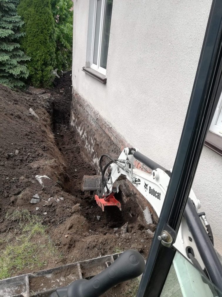 Mini koparka Bobcat wykonuje wąski wykop wzdłuż ściany budynku z widoczną podmurówką, ziemia usypana po bokach wykopu, drzewa iglaste w tle.