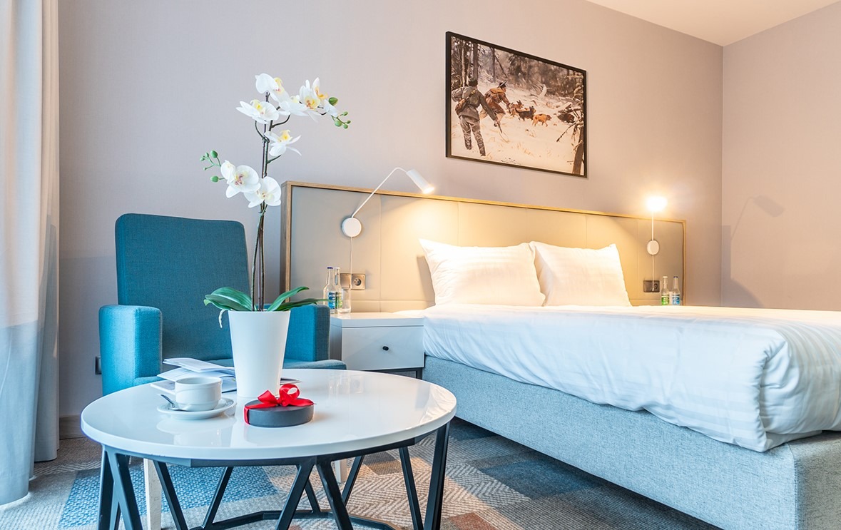 Nowoczesny pokój hotelowy z łóżkiem, turkusowym fotelem, okrągłym stolikiem z filiżanką i orchideą, oświetlony lampkami nocnymi i ozdobiony obrazem myśliwskim.