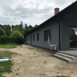 Jednopiętrowy dom z szarą elewacją w trakcie prac wykończeniowych, widoczne wystające rury instalacyjne na ścianie i niezabezpieczony teren wokół budynku.