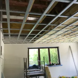 Wnętrze w trakcie montażu sufitu podwieszanego z widoczną konstrukcją metalową i częściowo ułożoną wełną mineralną, z rusztowaniem i materiałami budowlanymi w tle.