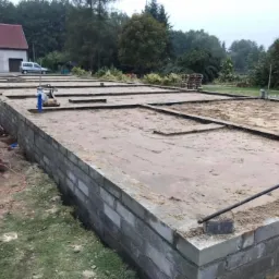 Wylany fundament z bloczków betonowych z widocznymi deskami szalunkowymi i przygotowanym podłożem z piasku, w tle dom jednorodzinny i zieleń.