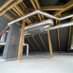 Instalacja systemu wentylacji z rekuperacją w surowym stanie poddasza, widoczne kanały wentylacyjne owinięte folią aluminiową, centrala wentylacyjna oraz drewniana konstrukcja dachu.