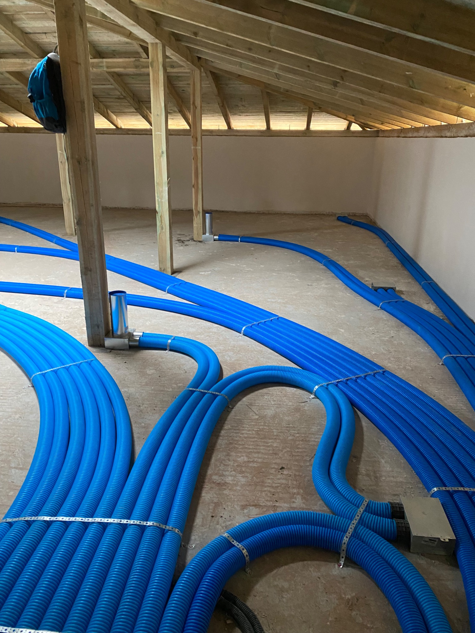 Niedokończona instalacja wentylacyjna na poddaszu: wiązki niebieskich, karbowanych przewodów wentylacyjnych przymocowane metalowymi taśmami, widoczne drewniane krokwie dachu i fragment ściany...