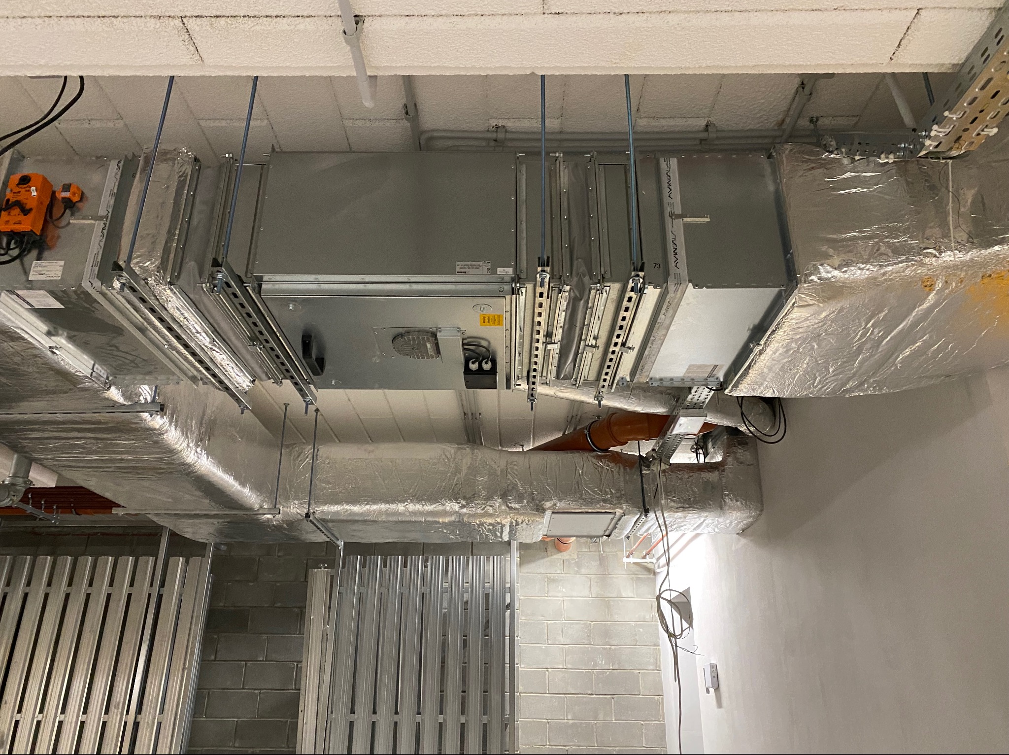 Widok na sufit z zamontowaną, srebrną instalacją kanałów wentylacyjnych, zabezpieczonych folią, z widocznym pomarańczowym siłownikiem i systemem podwieszeń na szynach montażowych.