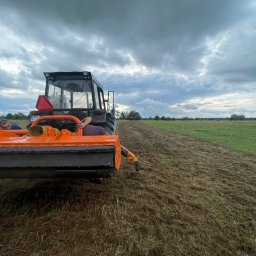 Alpina - Ciężki traktor z pomarańczowym mulczerem tnący trawę na polu w pochmurny dzień, widoczny fragment pola już po koszeniu.