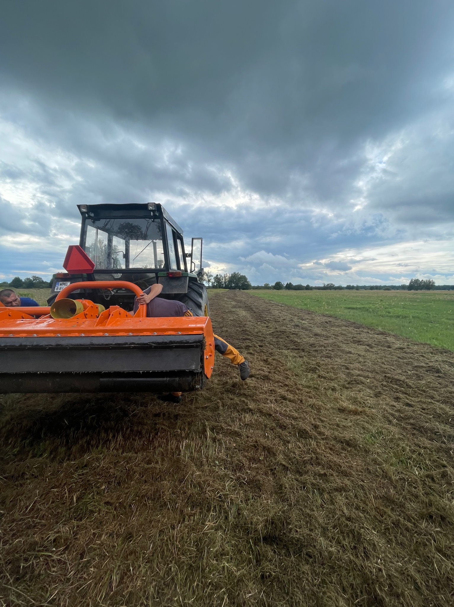 Ciężki traktor z pomarańczowym mulczerem tnący trawę na polu w pochmurny dzień, widoczny fragment pola już po koszeniu.