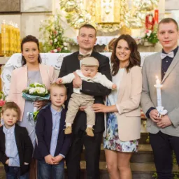 Uśmiechnięta rodzina z trzema synami pozuje w kościele podczas uroczystości, ojciec trzyma na rękach niemowlę w beżowej czapeczce.