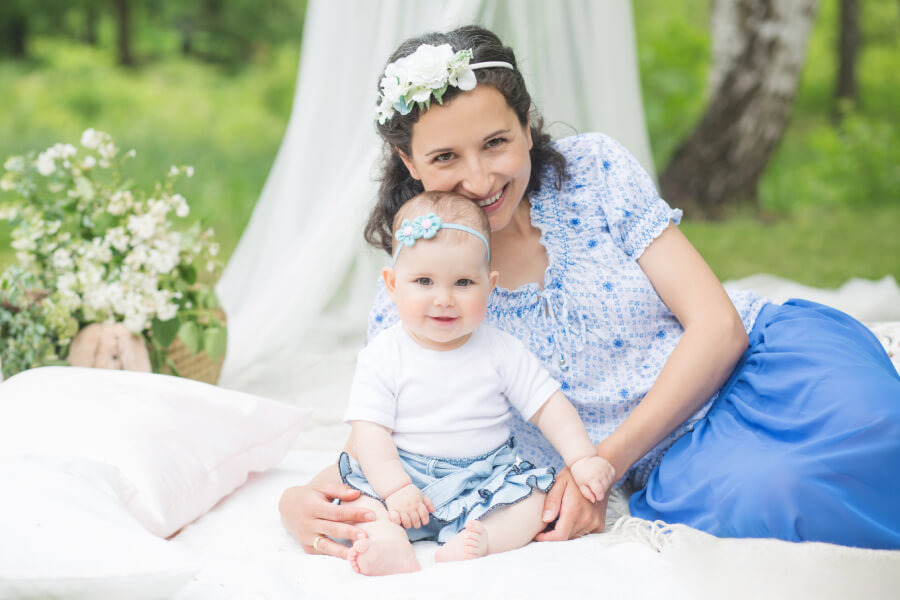 Uśmiechnięta mama z ciemnymi włosami i kwiatową opaską, trzymająca na kolanach uśmiechniętą córeczkę z błękitną opaską na głowie, podczas plenerowej sesji na białym kocu z kwiatami w tle.