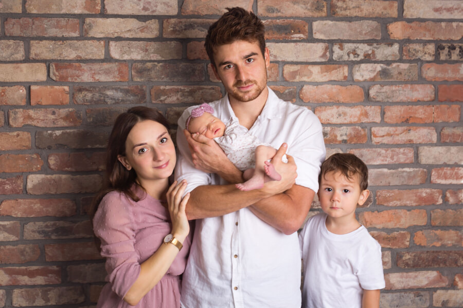 Rodzinne portret: tata trzyma śpiące niemowlę z fioletową opaską, mama w różowej sukience i syn w białej koszulce pozują na tle ceglanej ściany.