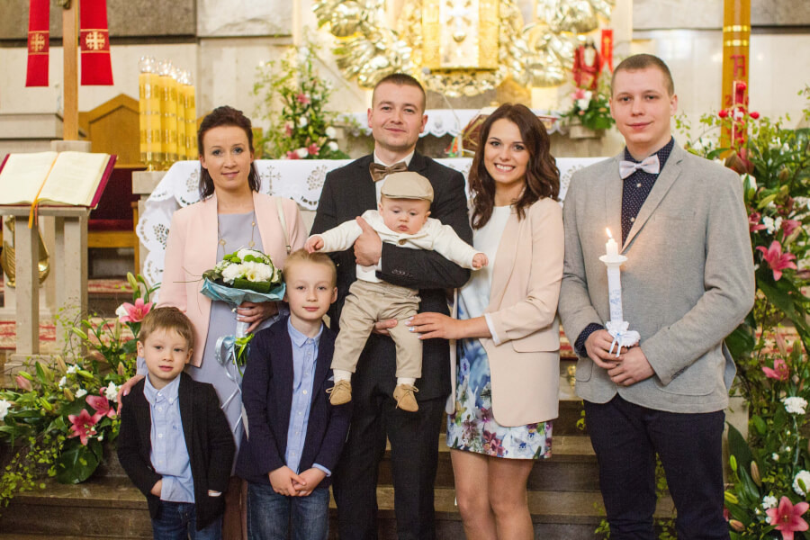 Uśmiechnięta rodzina z trzema synami pozuje w kościele podczas uroczystości, ojciec trzyma na rękach niemowlę w beżowej czapeczce.