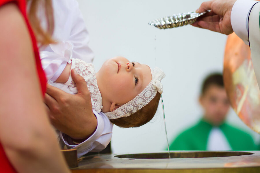 Dziecko w białej sukience i koronkowej opasce jest chrzczone wodą z srebrnej miseczki, w tle widoczna osoba w zielonej szacie.