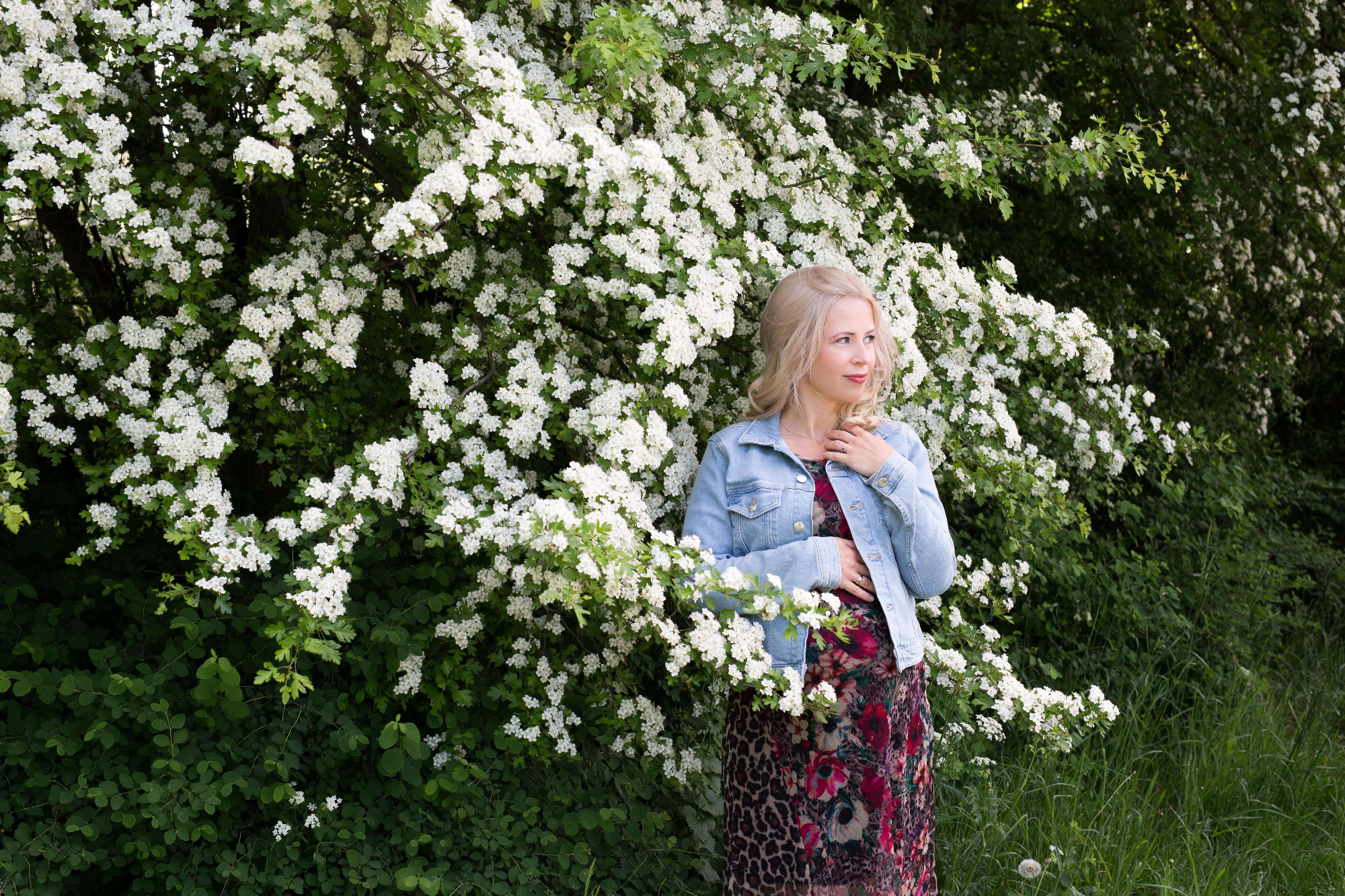 Kobieta w kwiecistej sukience i dżinsowej kurtce pozuje na tle kwitnącego krzewu z białymi kwiatami, trzymając rękę na brzuchu.