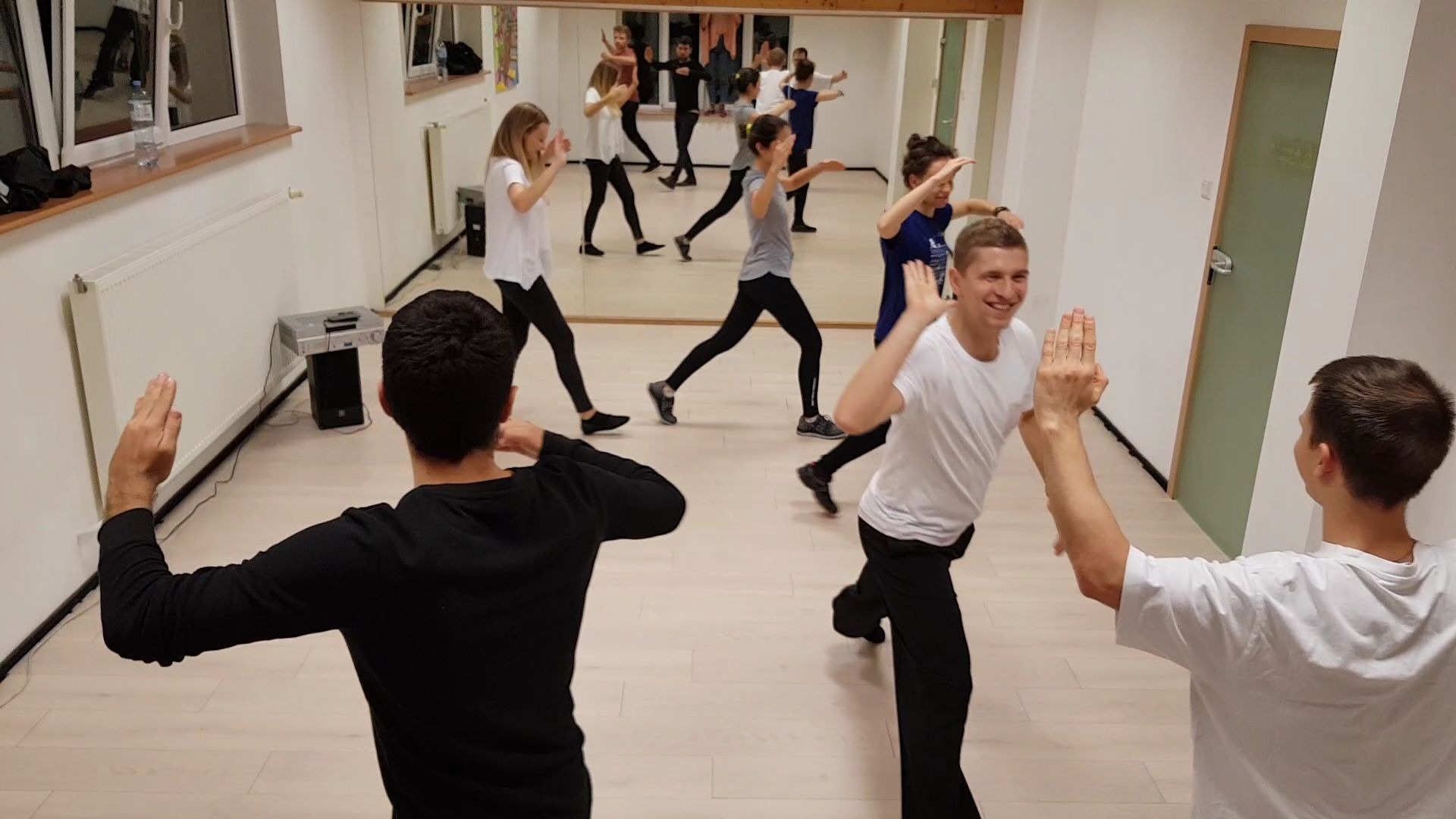 Grupa osób w jasnej sali, ćwiczących choreografię taneczną, widocznych w lustrze na ścianie.