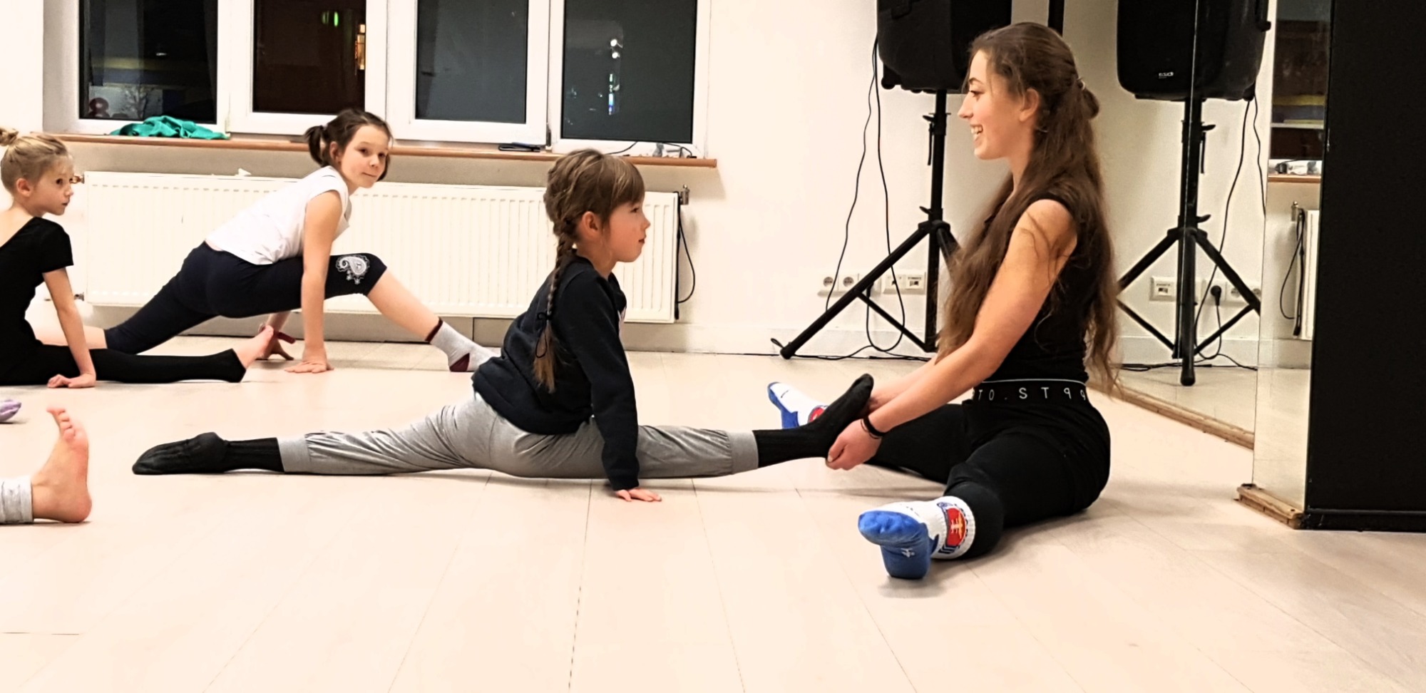 Instruktorka pomaga dziewczynce w szpagacie podczas zajęć w szkole tańca, inne dzieci ćwiczą w tle, widoczne okno i głośnik.