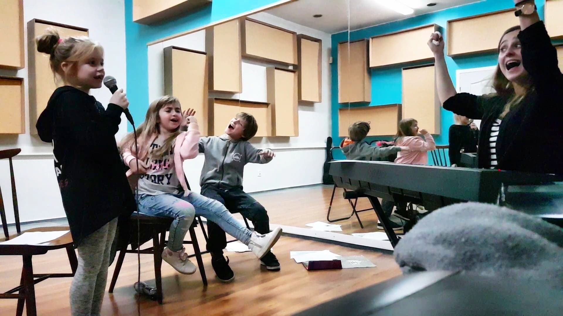 Młoda dziewczynka śpiewa do mikrofonu, siedzący obok chłopiec i dziewczynka z entuzjazmem gestykulują, nauczycielka gra na keyboardzie i podnosi ręce w górę, w tle panele akustyczne na ścianie.