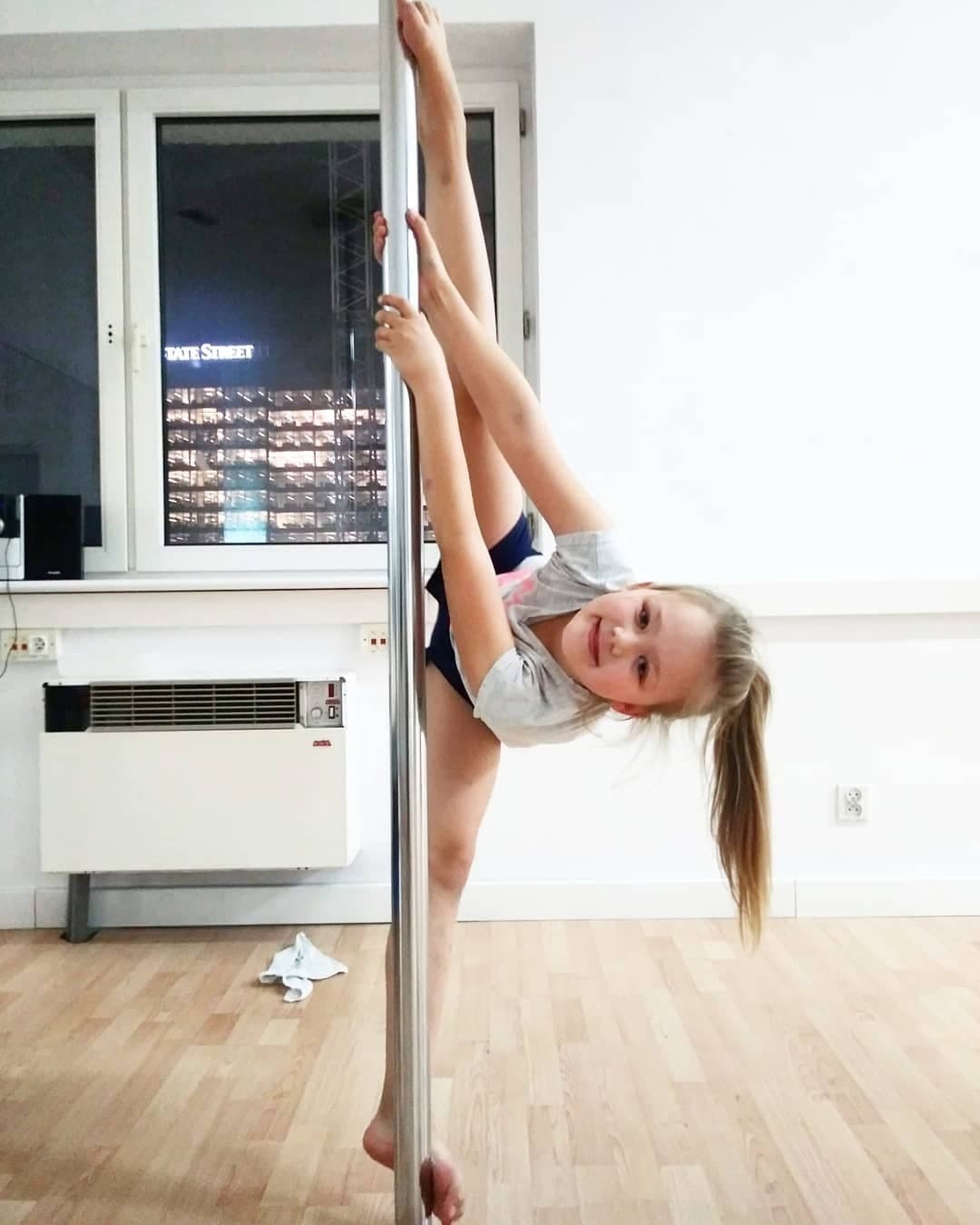Dziewczynka uśmiecha się, wykonując figurę na rurze w jasnym pomieszczeniu z drewnianą podłogą i oknem z widokiem na nocne miasto.