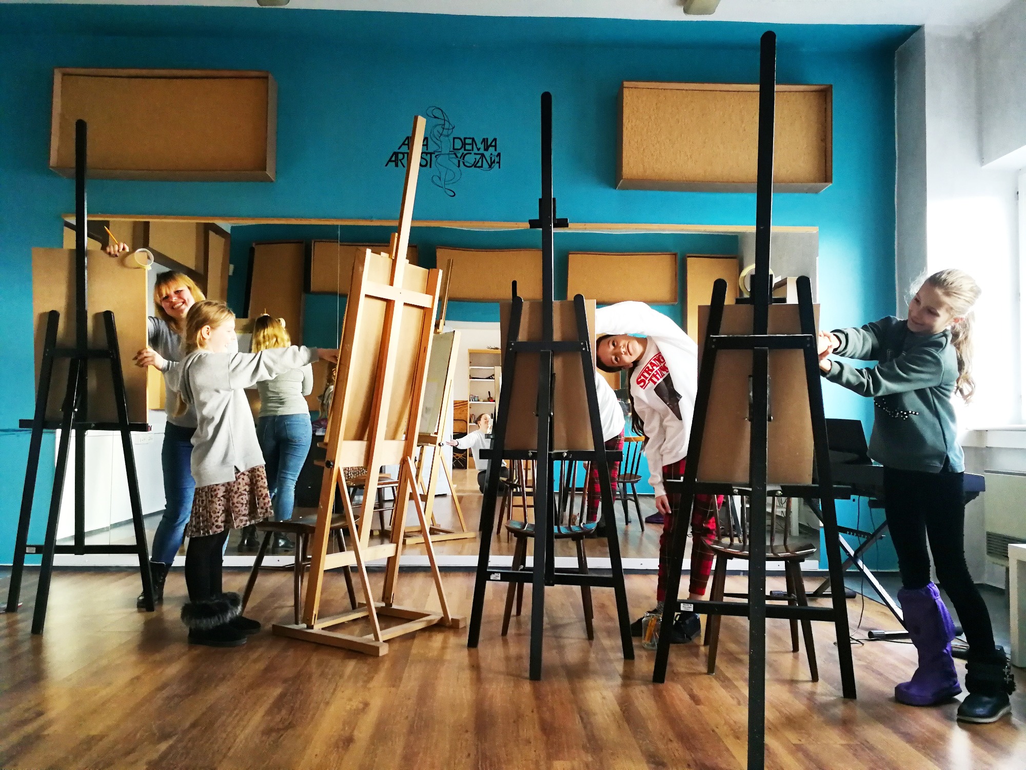 Grupa dzieci i dorosłych w pracowni artystycznej, malujący przy sztalugach, widoczne obrazy, lustro i napis 'Akademia Artystyczna' na ścianie.
