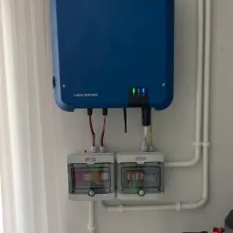 Niebieski falownik SMA Sunny Tripower zamontowany na białej ścianie, z widocznymi przewodami i dwoma zabezpieczeniami ETI, obok białych rur instalacyjnych i fragmentu zbiornika z zaworem.