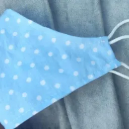 Jasnoniebieska maseczka ochronna w białe kropki, uszyta w Bedlnie, z białymi elastycznymi gumkami, leżąca na szarym tle.