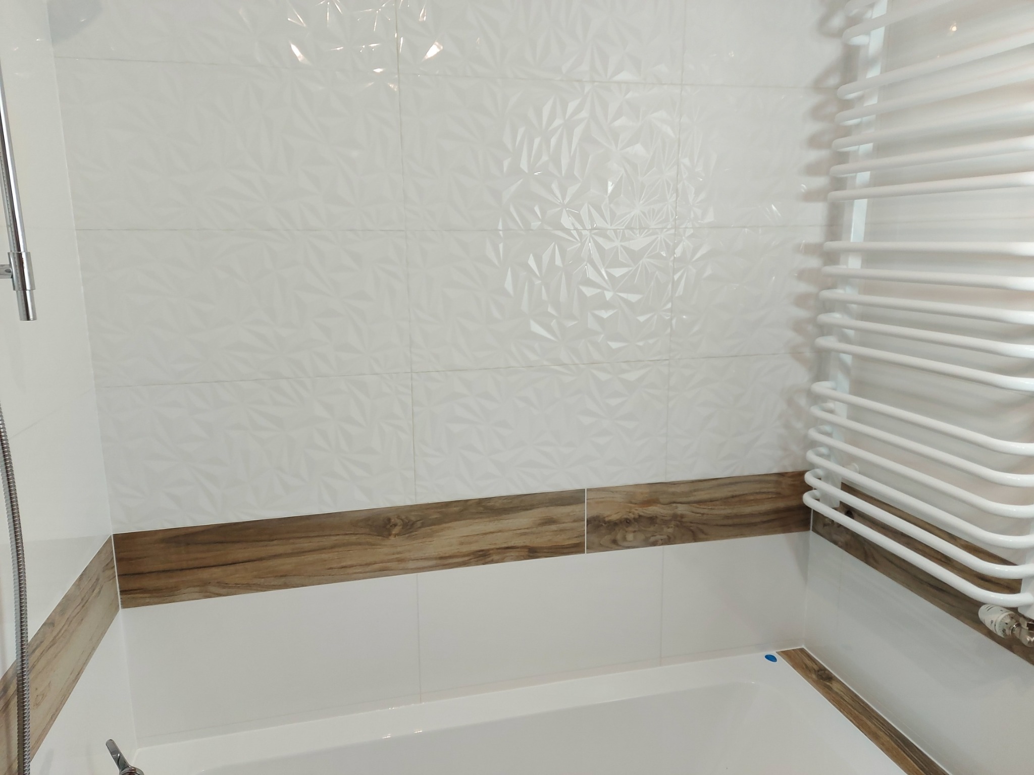 Białe płytki 3D na ścianie łazienki z drewnopodobnym pasem dekoracyjnym i grzejnikiem drabinkowym.