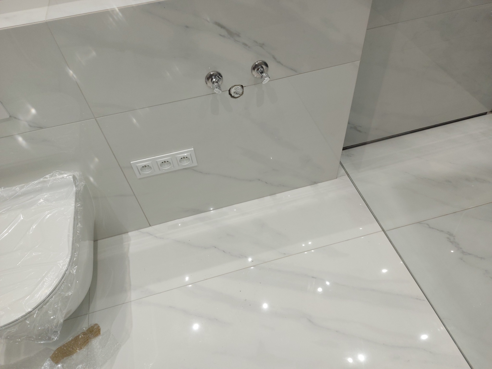 Białe płytki imitujące marmur na ścianie i podłodze w łazience, z widocznymi chromowanymi zaworami i gniazdkami elektrycznymi, częściowo zakryta folią ochronną biała miska WC.