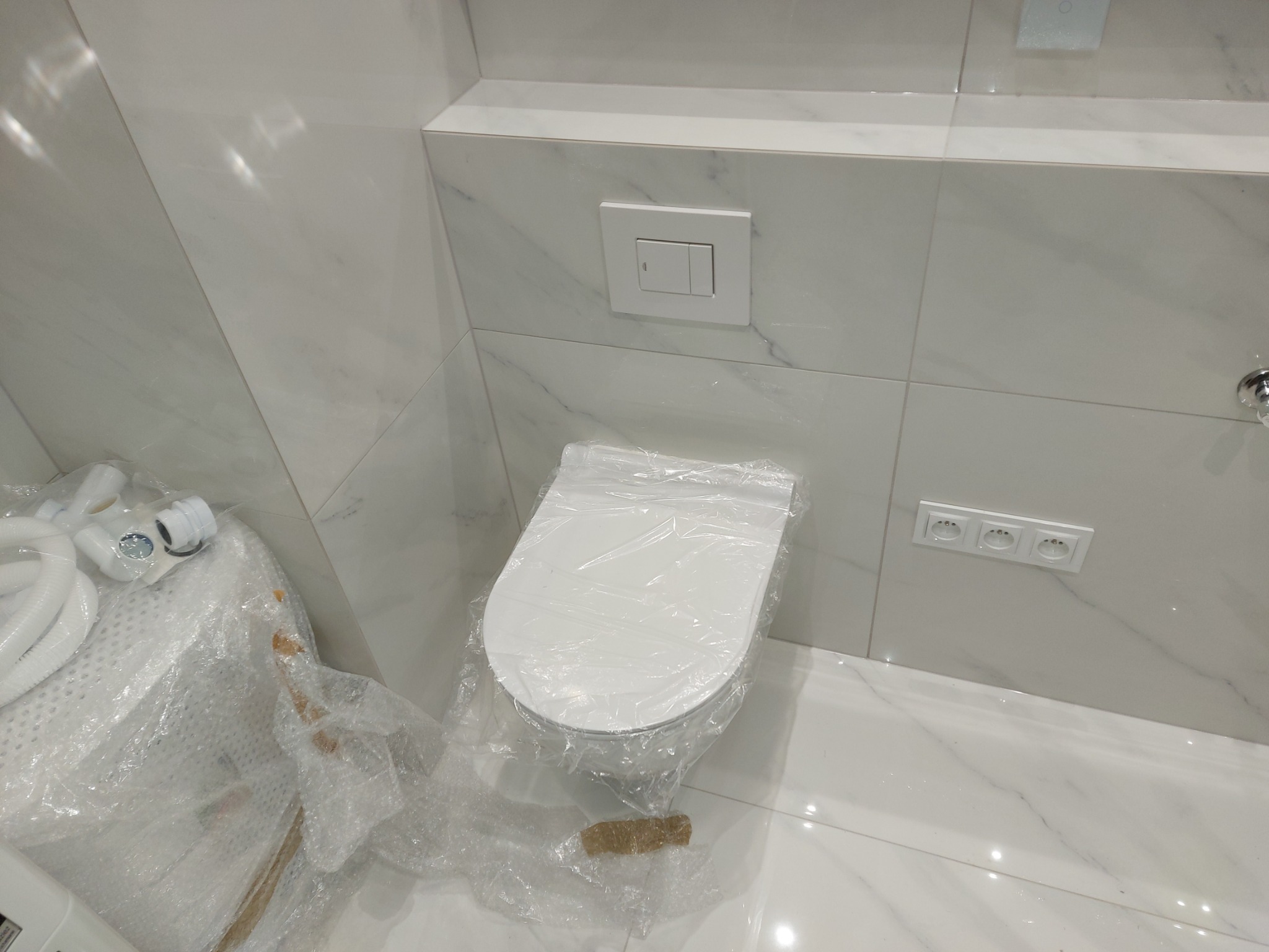 Nowa, biała toaleta w folii ochronnej, zamontowana w łazience wykończonej marmurowymi płytkami, widoczne gniazdka elektryczne i przycisk spłuczki w Warszawie.