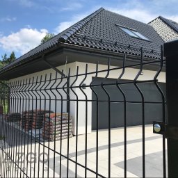 AZGO SP. Z O.O. - Nowoczesne, czarne ogrodzenie segmentowe z charakterystycznym falistym wykończeniem, przed białym domem z czarnym dachem i bramą garażową. W tle worki z materiałami ogrodniczymi.