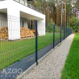 AZGO SP. Z O.O. - Ciemnozielony płot panelowy z betonową podmurówką, oddzielający nowoczesny dom z ułożonym drewnem kominkowym od trawnika i żwirowej ścieżki, z lasem w tle.