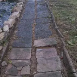 Ścieżka z nieregularnych kamiennych płyt prowadząca przez trawnik, z widocznym brzegiem trawnika i fragmentem oczka wodnego z kamieniami na obrzeżach, zarośnięta trawa między płytami.