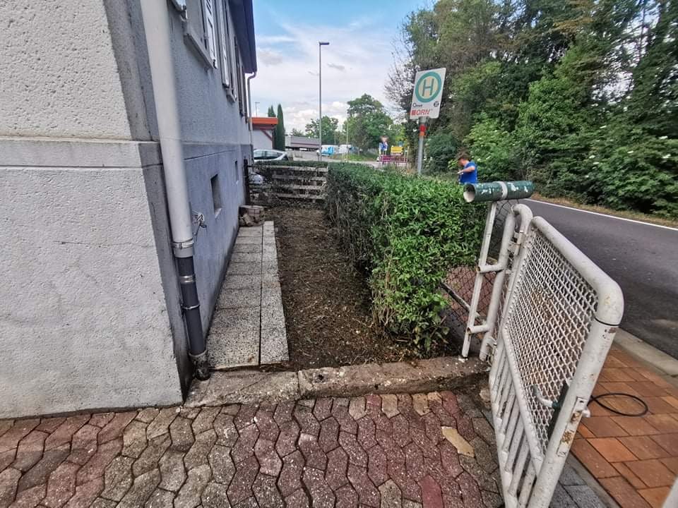 Przygotowanie terenu pod nasadzenia wzdłuż ściany budynku, widoczna rabata z ziemią, przycięty żywopłot i tymczasowe ogrodzenie zabezpieczające.