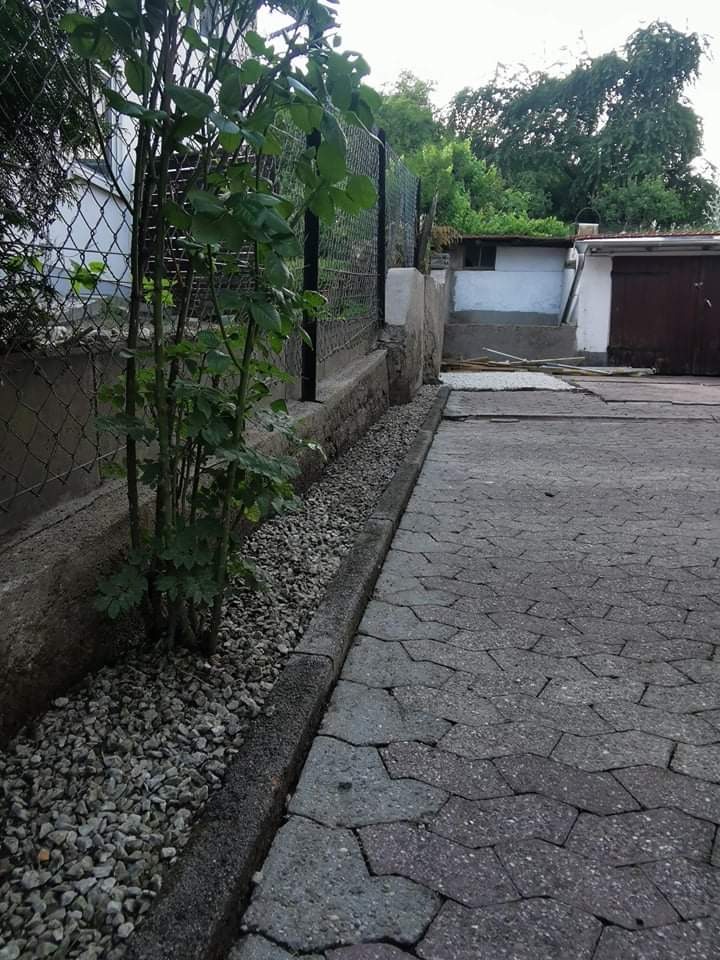 Ułożona kostka brukowa wzdłuż ogrodzenia z nasadzeniami i drobnym żwirem, widoczny garaż w tle.