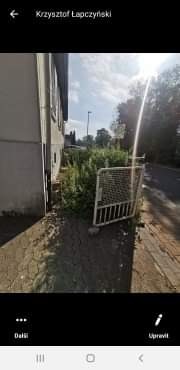 Zarośnięty ogródek przed domem z uchyloną, metalową furtką, widok z chodnika.