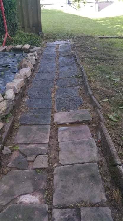 Ścieżka z nieregularnych kamiennych płyt prowadząca przez trawnik, z widocznym brzegiem trawnika i fragmentem oczka wodnego z kamieniami na obrzeżach, zarośnięta trawa między płytami.