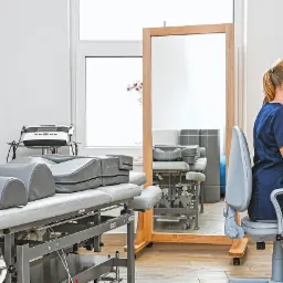 Fizjoterapeutka w gabinecie, siedząca przy biurku i pracująca na komputerze, w tle widoczny stół do terapii manualnej i lustro.