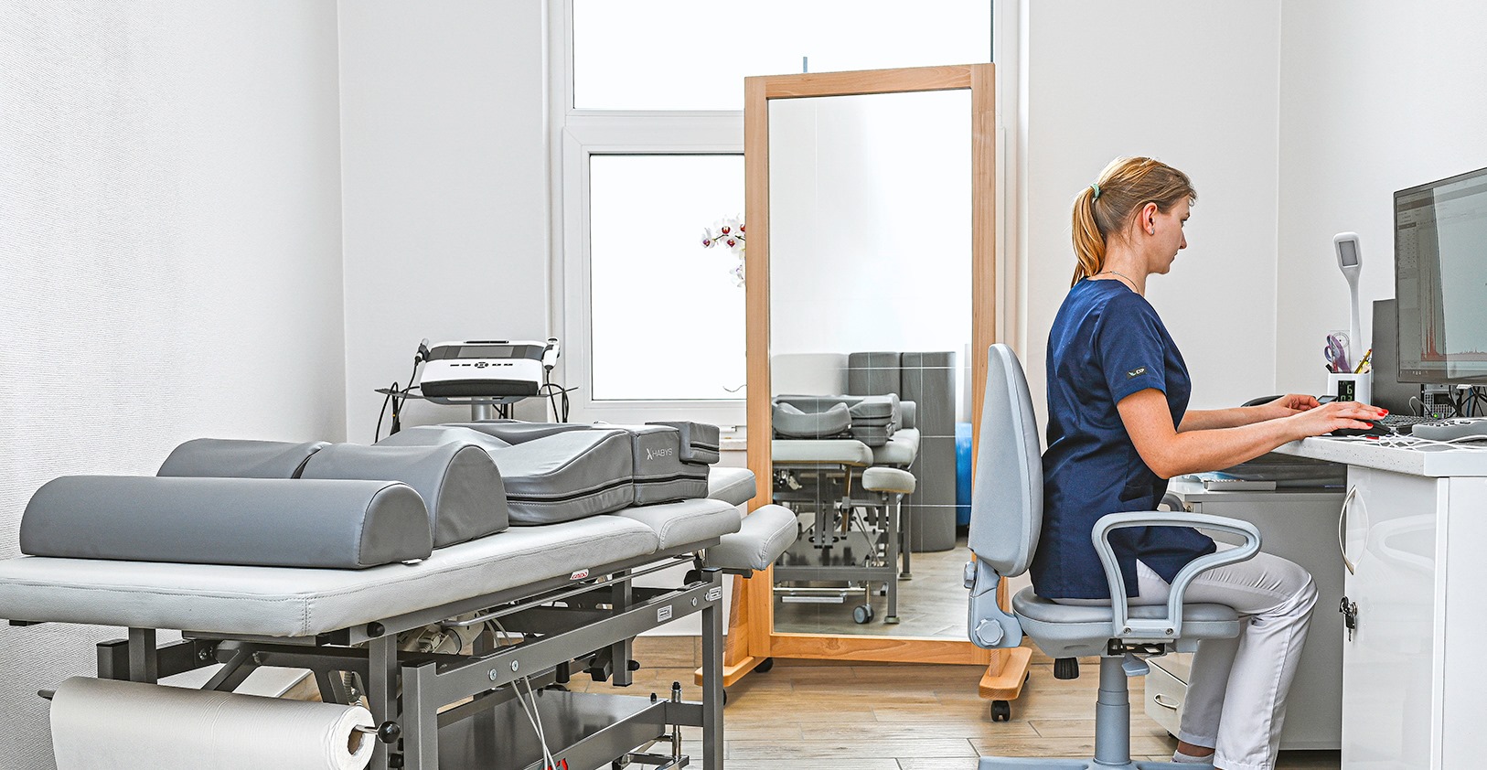 Fizjoterapeutka w gabinecie, siedząca przy biurku i pracująca na komputerze, w tle widoczny stół do terapii manualnej i lustro.