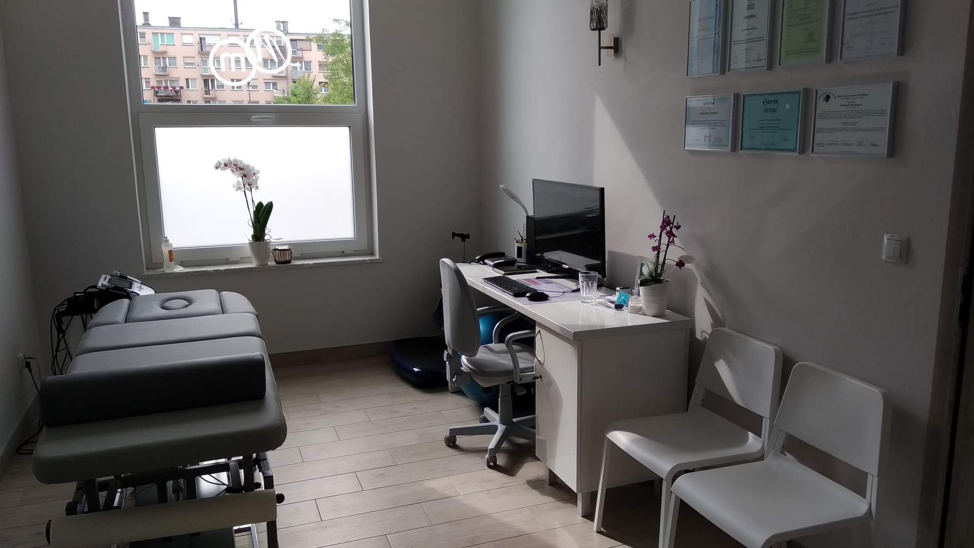 Jasne wnętrze gabinetu z leżanką do zabiegów, biurkiem z komputerem i krzesłami dla pacjentów. Na ścianie wiszą certyfikaty. Za oknem widoczny budynek mieszkalny.