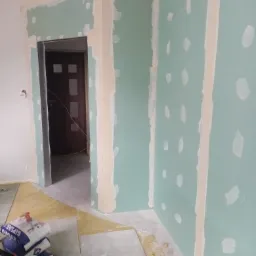 Wnętrze w trakcie prac wykończeniowych: ściany pokryte płytami gipsowo-kartonowymi, z widocznymi łączeniami i śladami szpachlowania, otwarte przejście drzwiowe, worki z zaprawą gipsową i narzędzia...