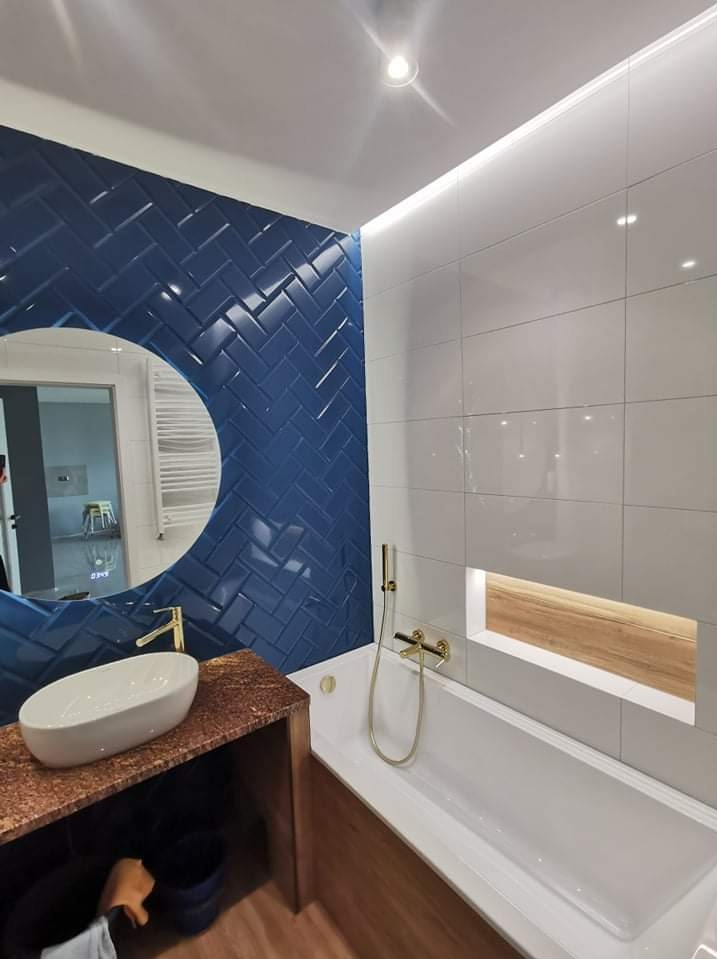 Nowoczesna łazienka z niebieskimi płytkami ułożonymi w jodełkę, okrągłym lustrem, białą umywalką na blacie z brązowego kamienia, białą wanną i złotą armaturą, oświetleniem LED pod sufitem i w niszy...