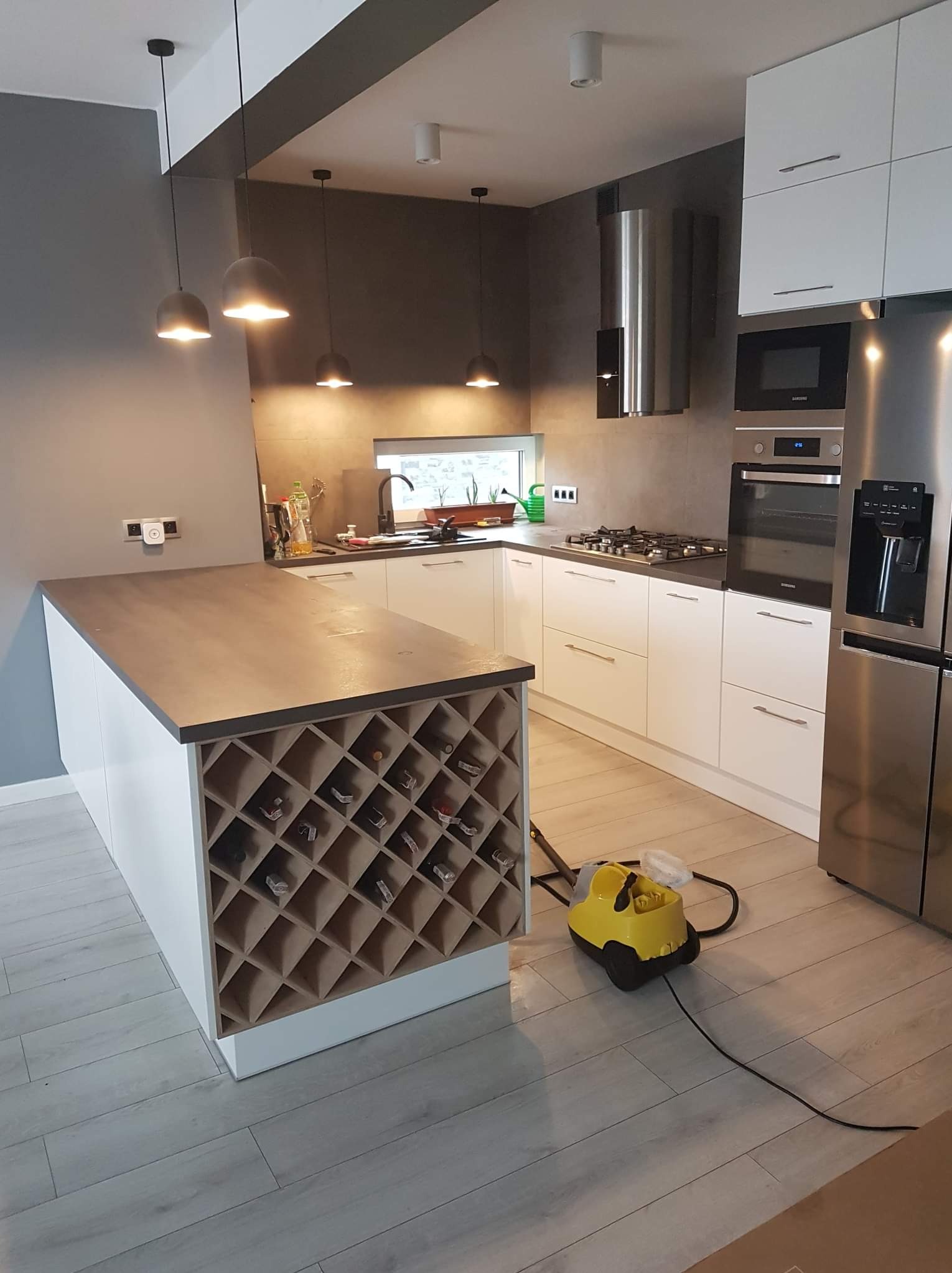 Nowoczesna kuchnia z wyspą i regałem na wino, oświetlona wiszącymi lampami, z żółtym parownikiem do czyszczenia podłogi na drewnianych panelach.